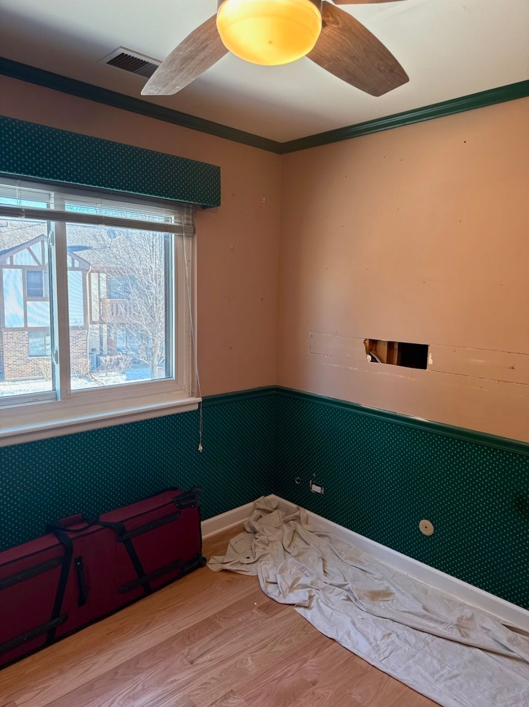Room with peach and green walls, window, ceiling fan, torn sheet on floor, and hole in wall.