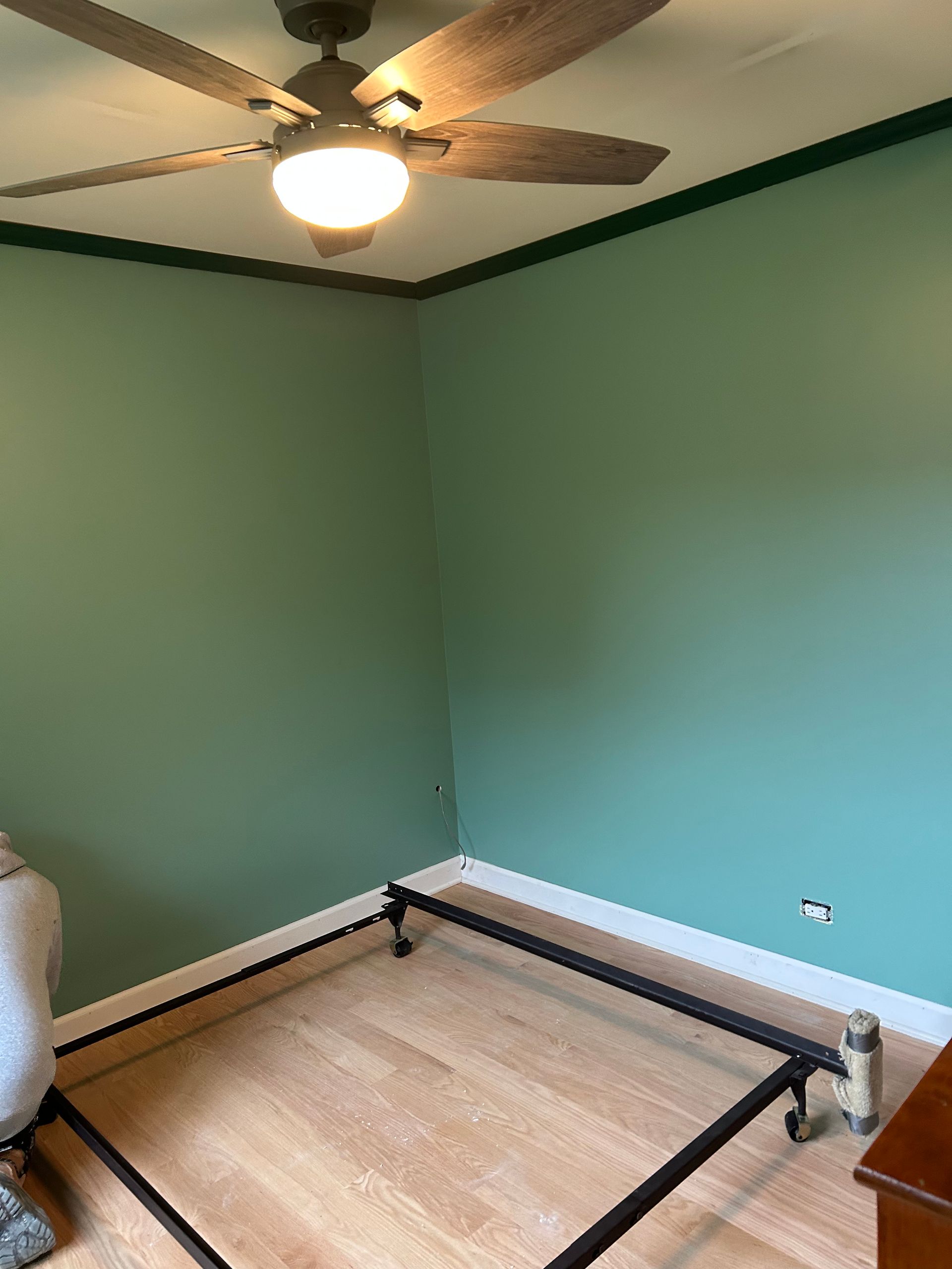 Empty bedroom with green walls, ceiling fan, bed frame, and wood floor.