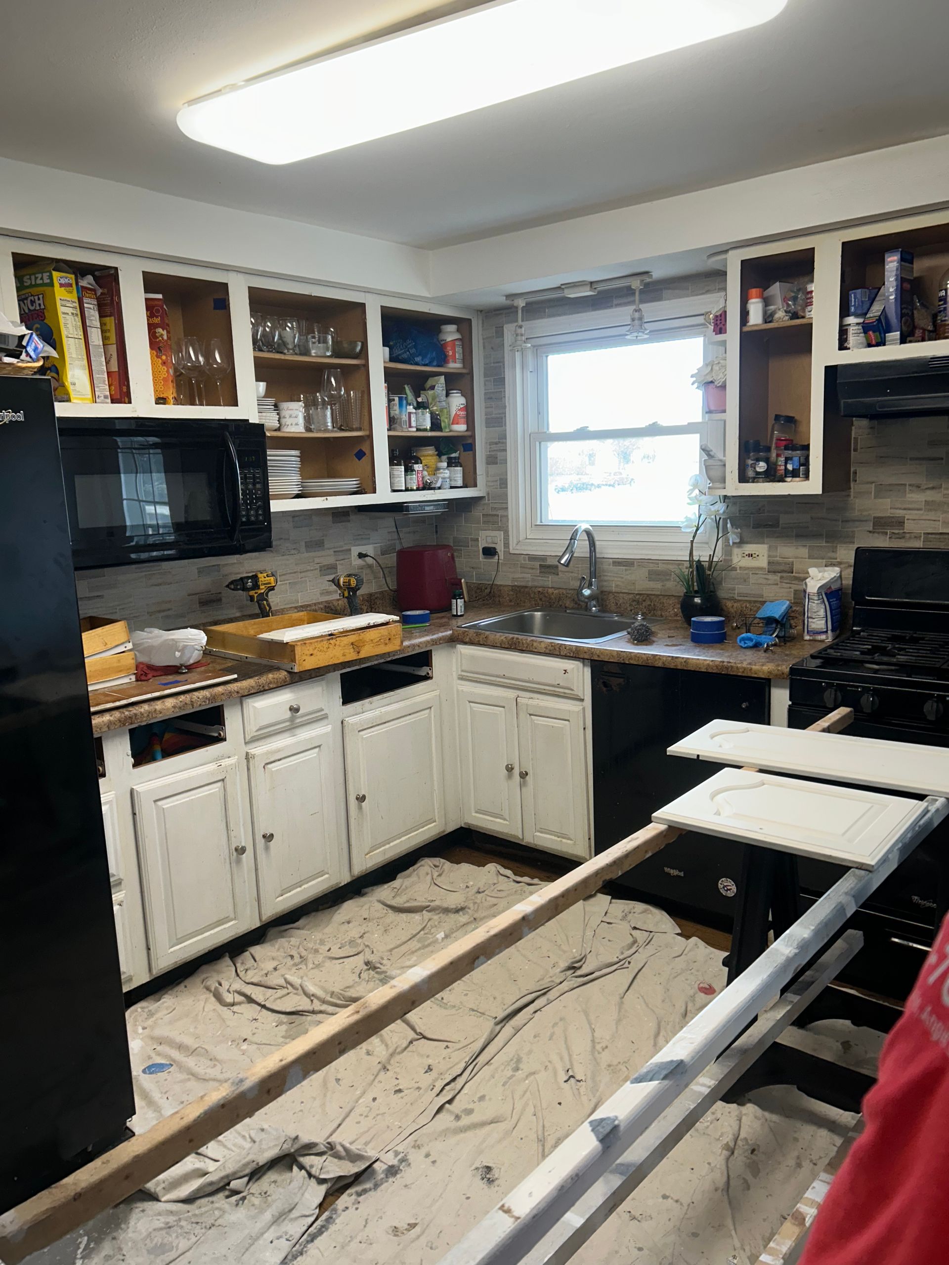 Kitchen renovation in progress, white cabinets, black appliances, window, painting supplies, paint on the floor.