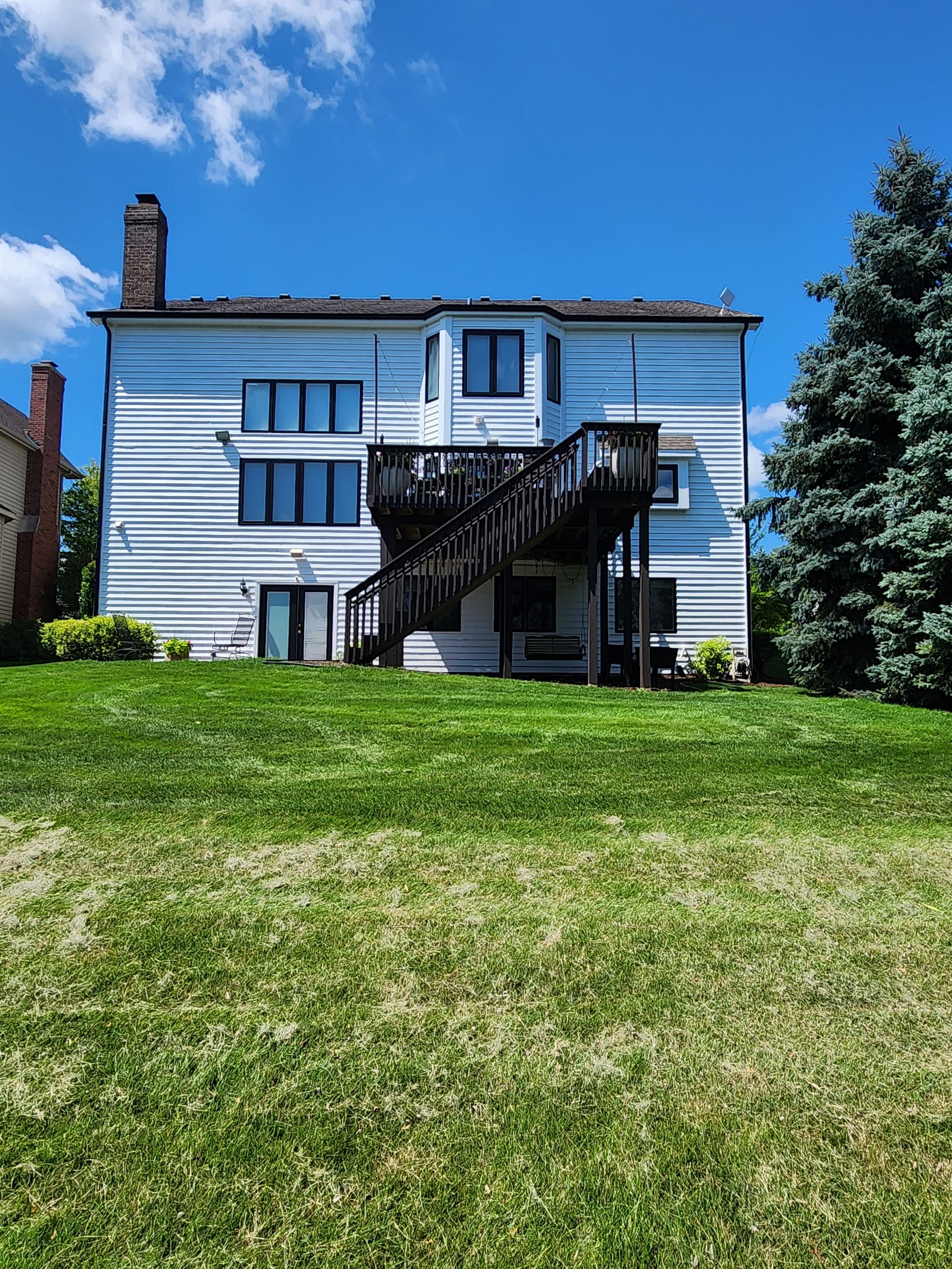 A large white house with a deck is sitting on top of a lush green hill.