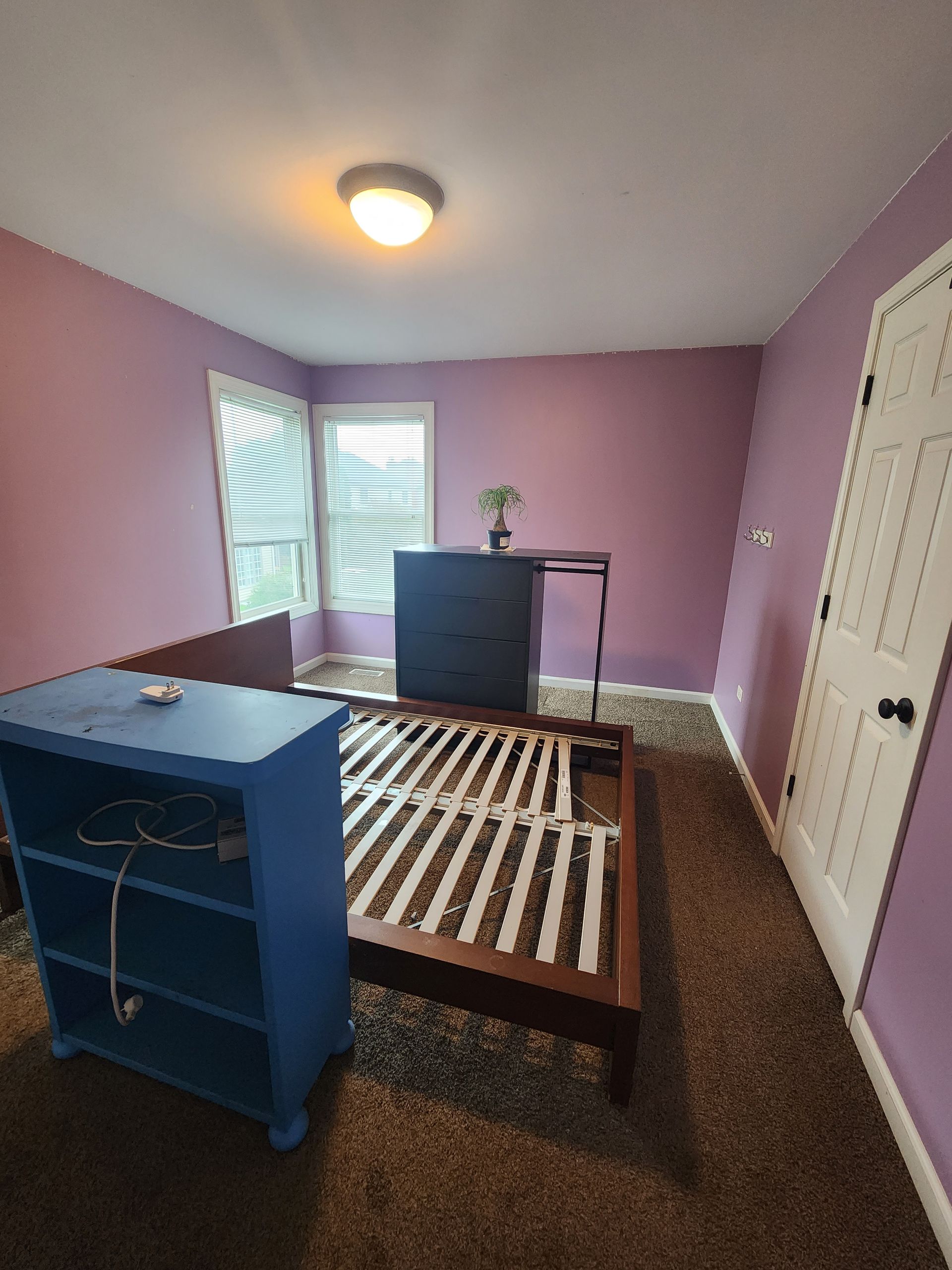 A bedroom with purple walls , a bed , a dresser and a blue shelf.