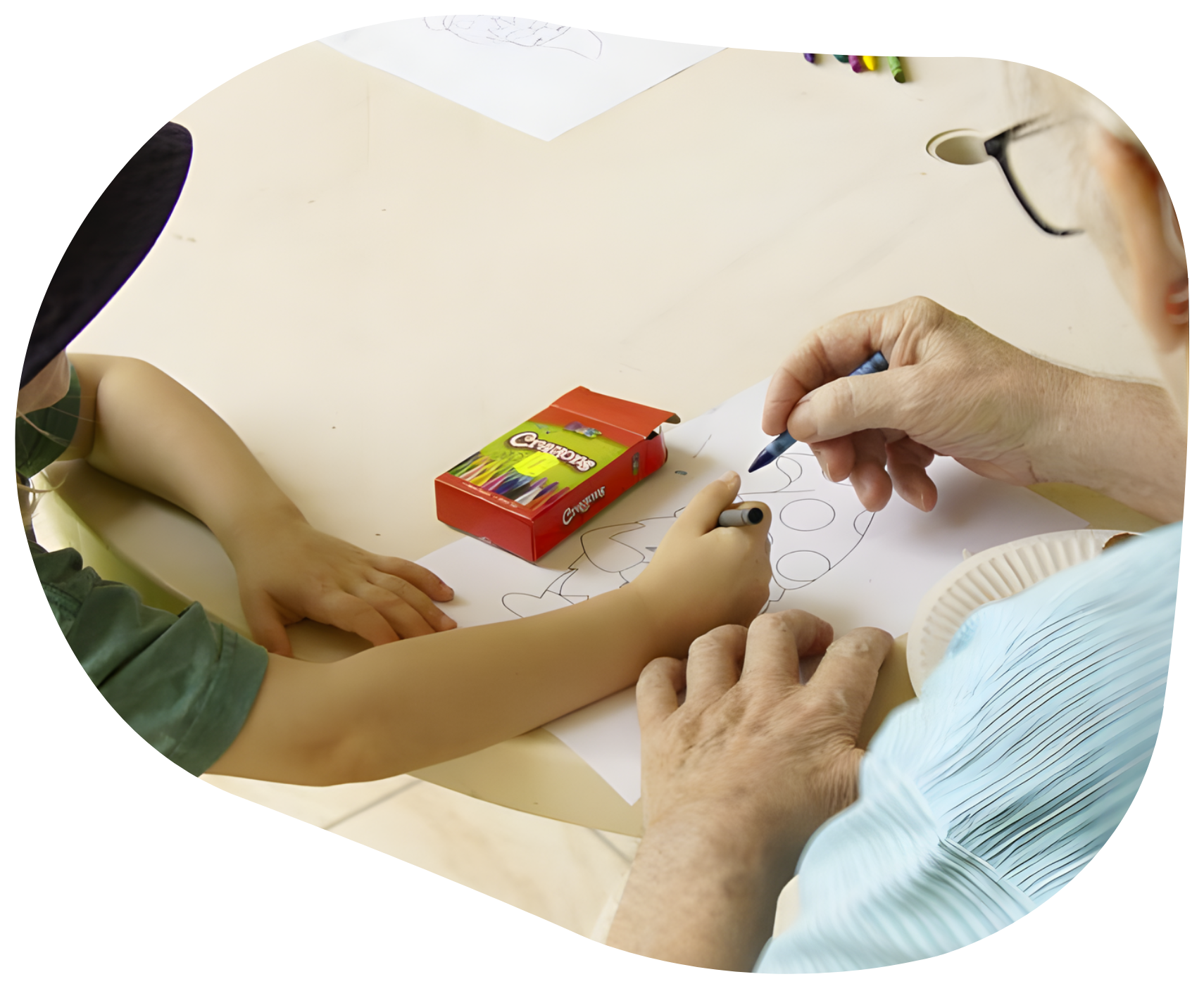 A child and adult are drawing together at a table. The adult's hand guides the child's hand with a pen.
