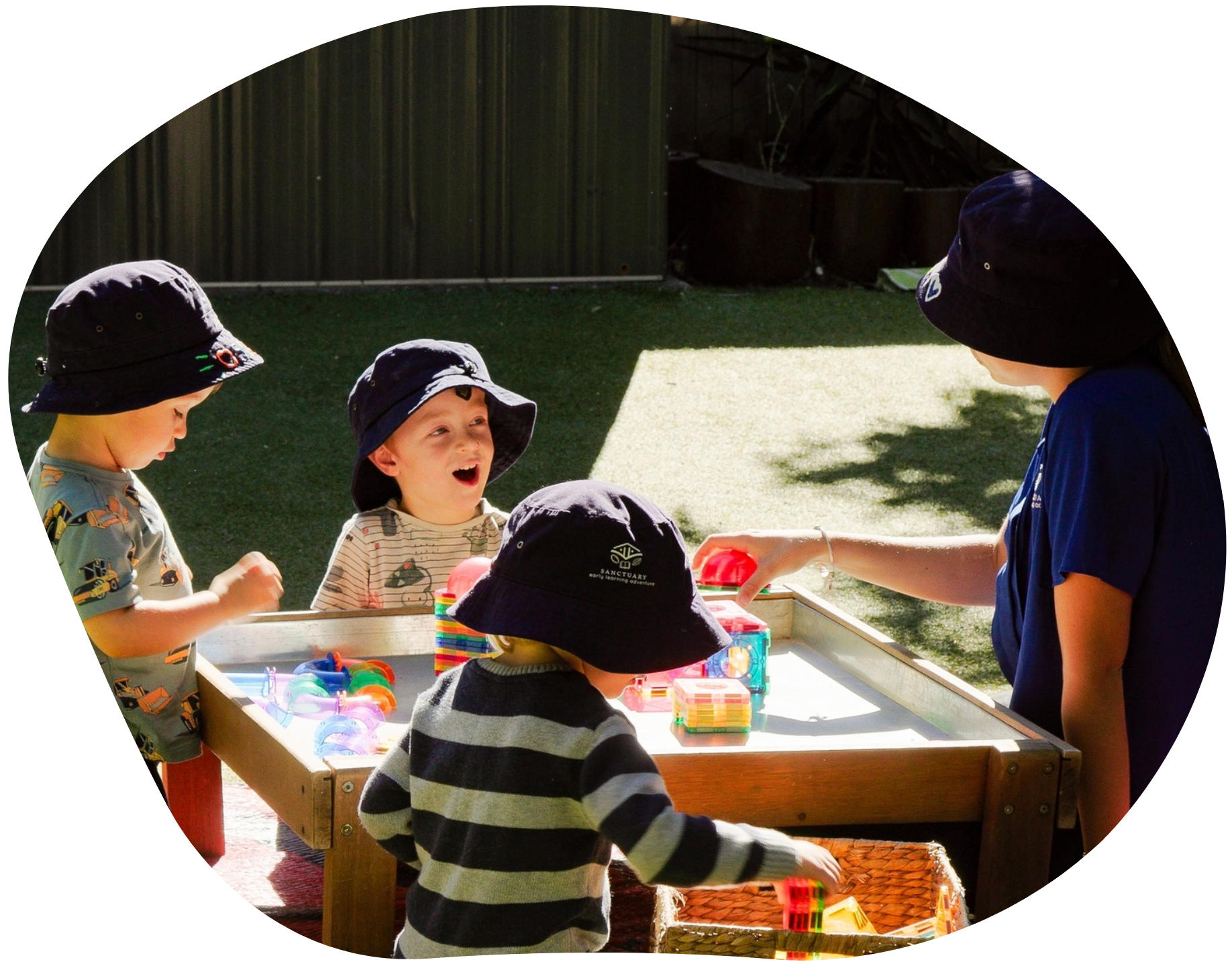 Children at a water table with a teacher outdoors. The children wear hats and play with blocks.