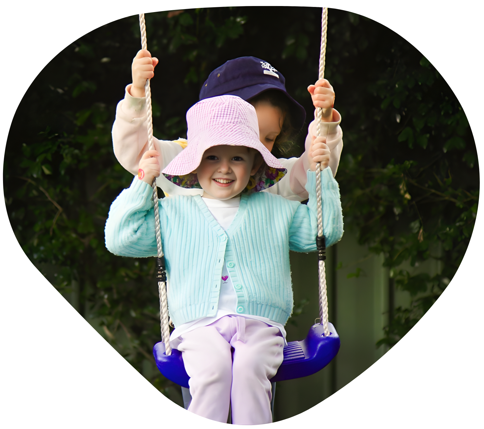 Two children on a swing. One in a blue sweater smiles, the other in a hat holds the ropes.