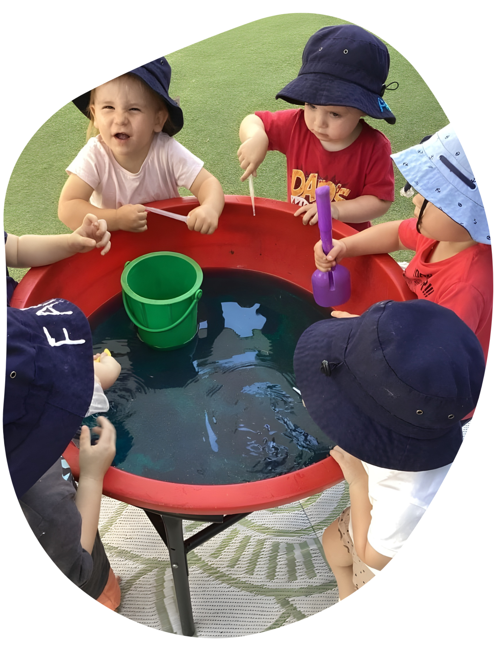 Children in hats play with water and toys in a red tub outdoors.