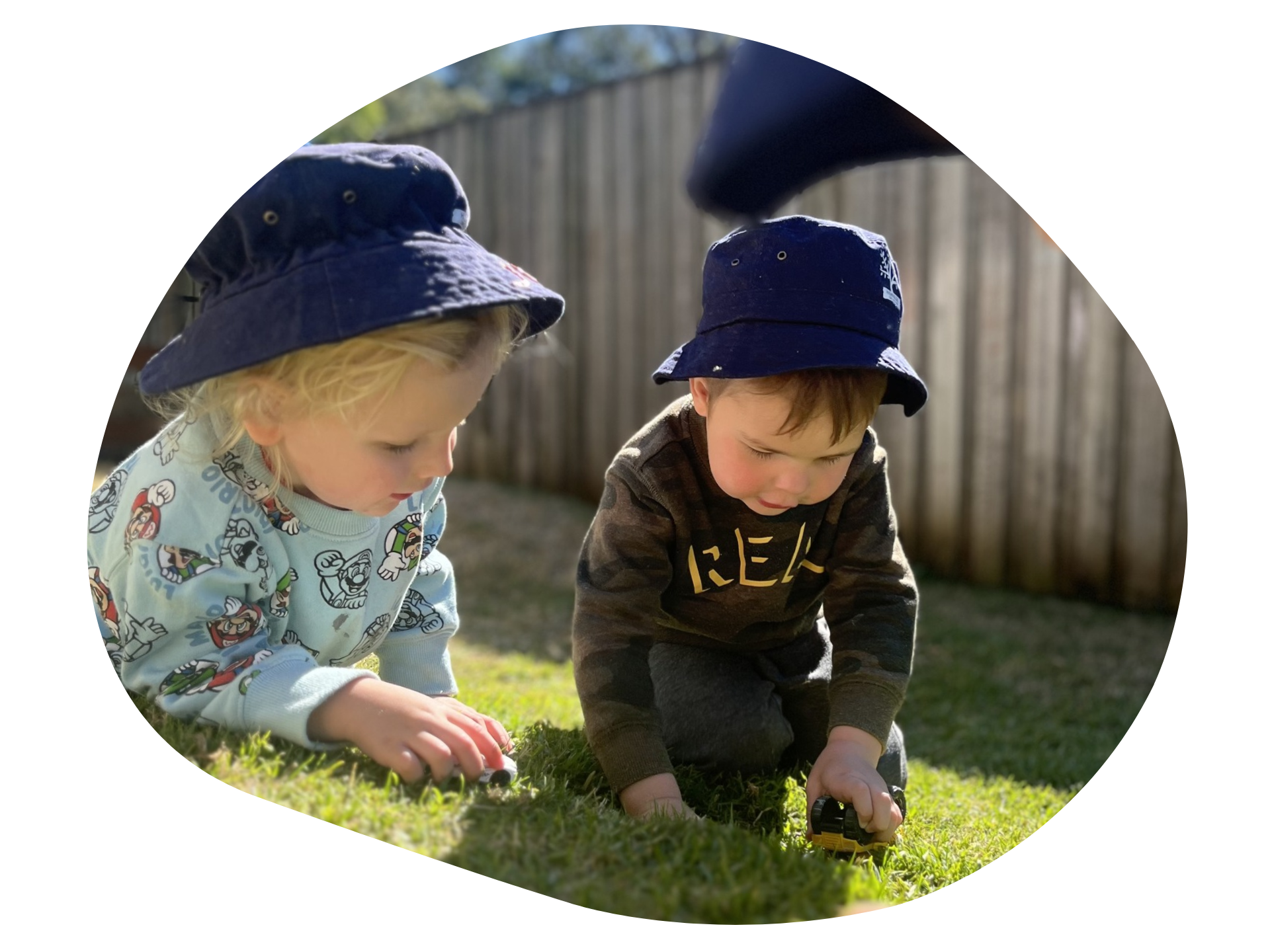 Two young children playing with toys on grass, wearing blue bucket hats.