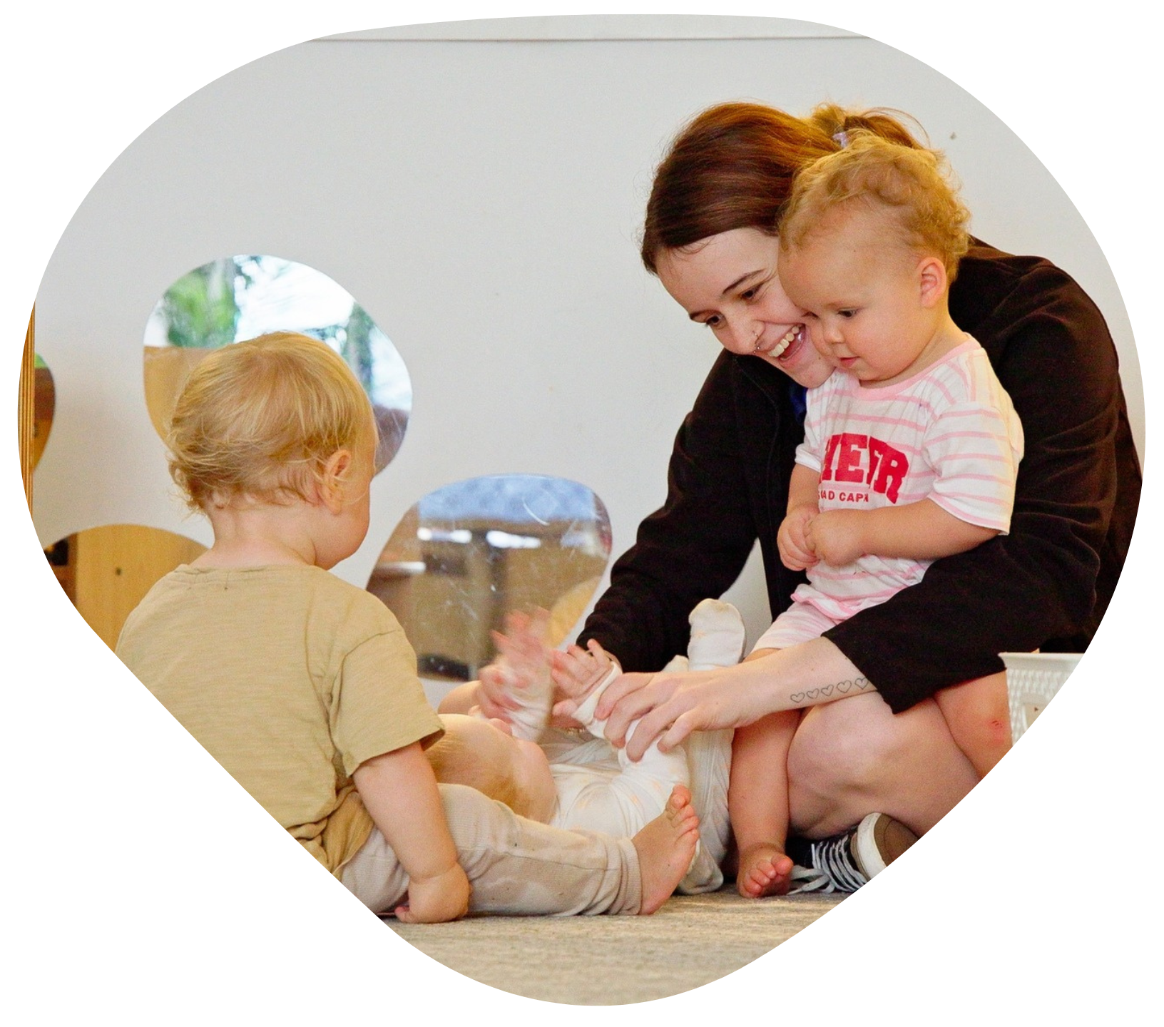 Woman changing a baby's diaper, another toddler watches. White room, soft colors.