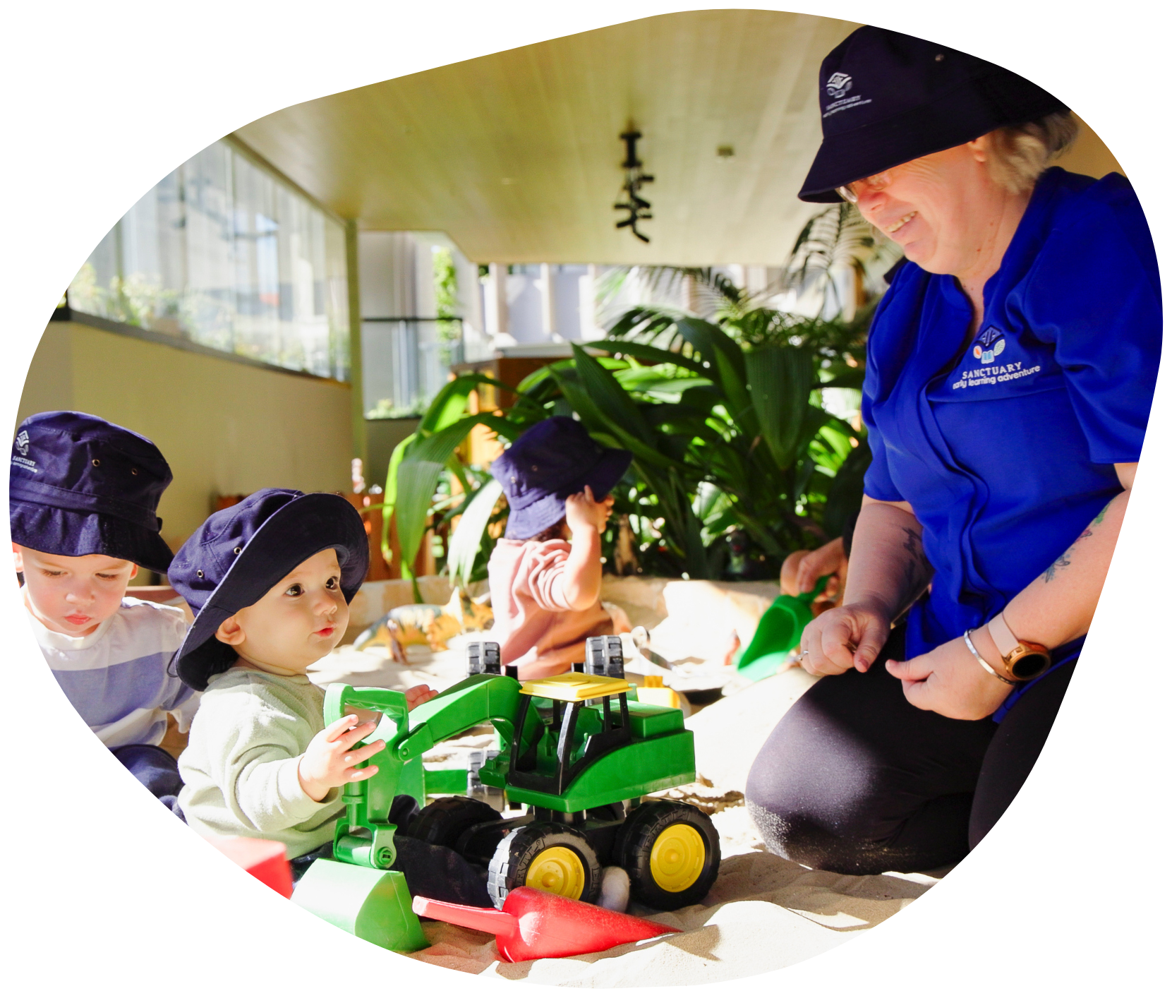Children in hats playing with a toy tractor and a teacher in an outdoor area.
