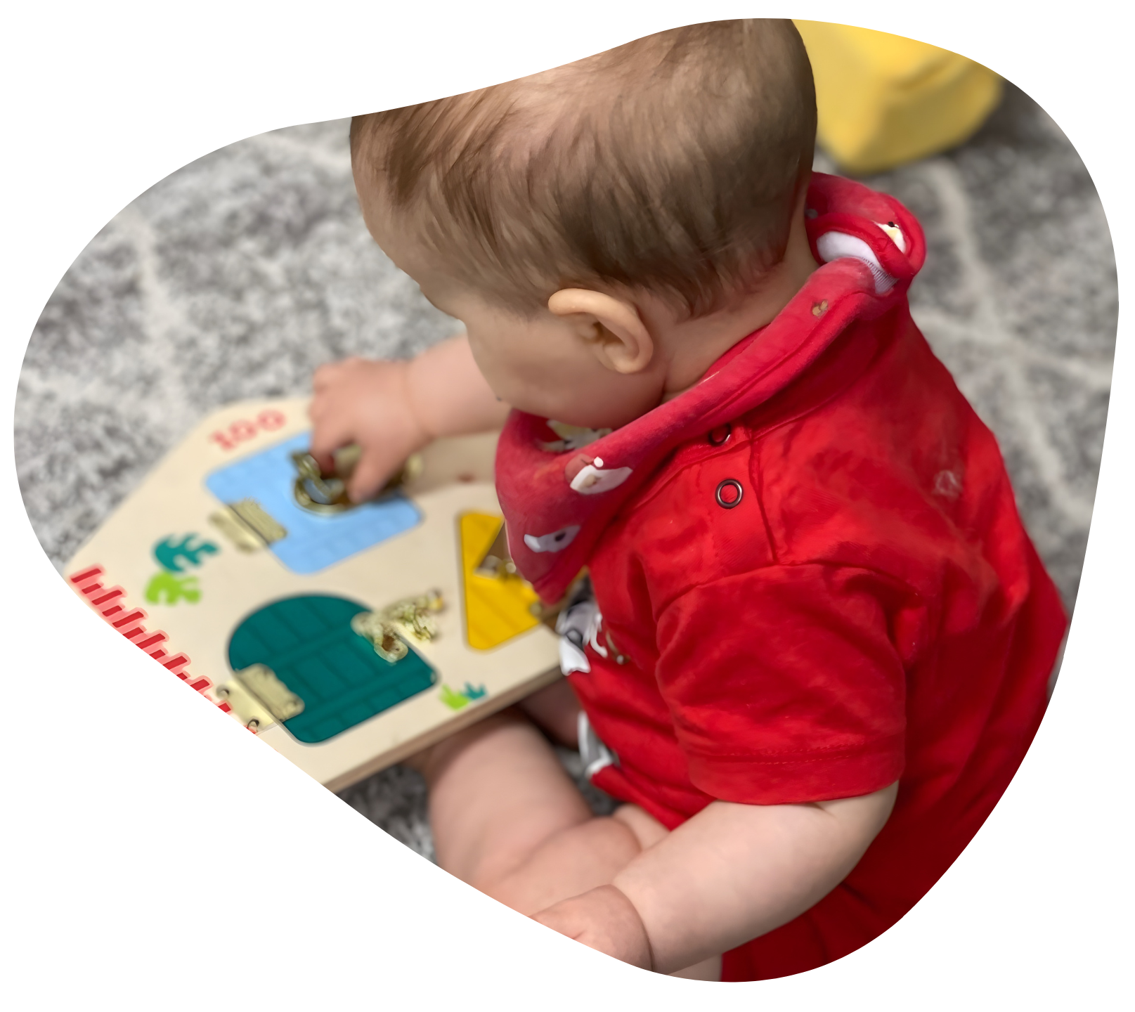 Baby in red shirt playing with a wooden toy puzzle on a gray rug.
