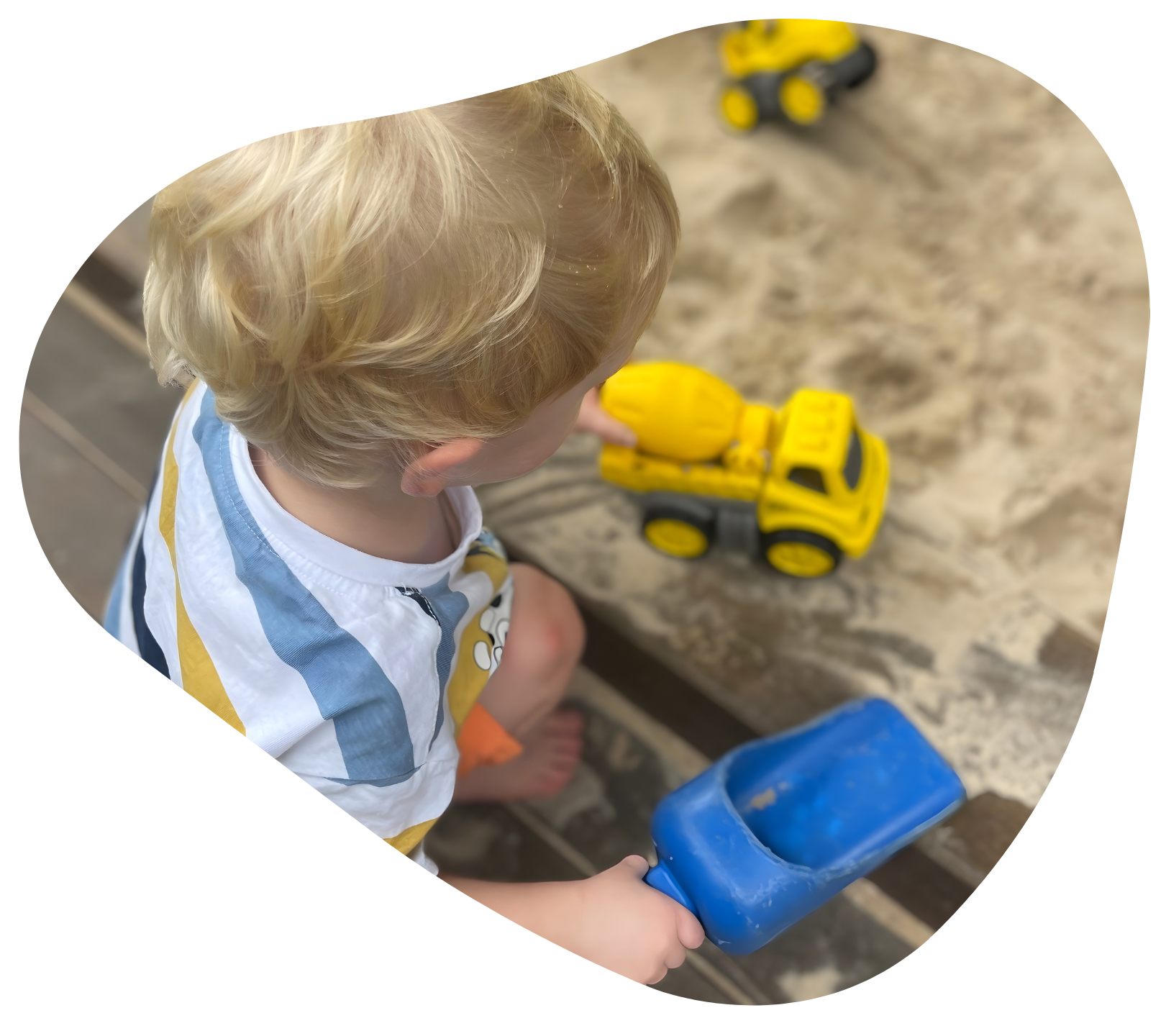 Child in striped shirt playing in sandbox with yellow truck and blue scoop.