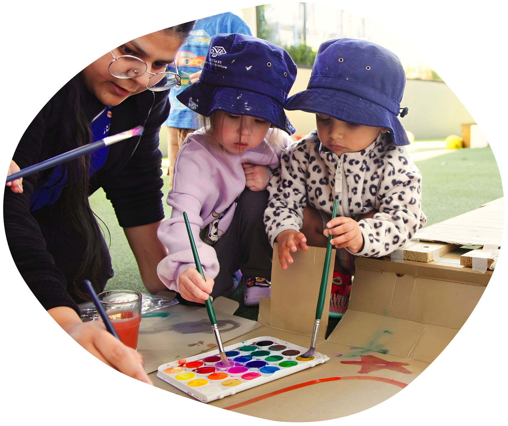 Children in blue hats paint with watercolors with an adult, outdoors.