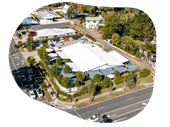 Aerial view of a commercial building with a white roof, surrounded by trees and a parking lot, near a road.