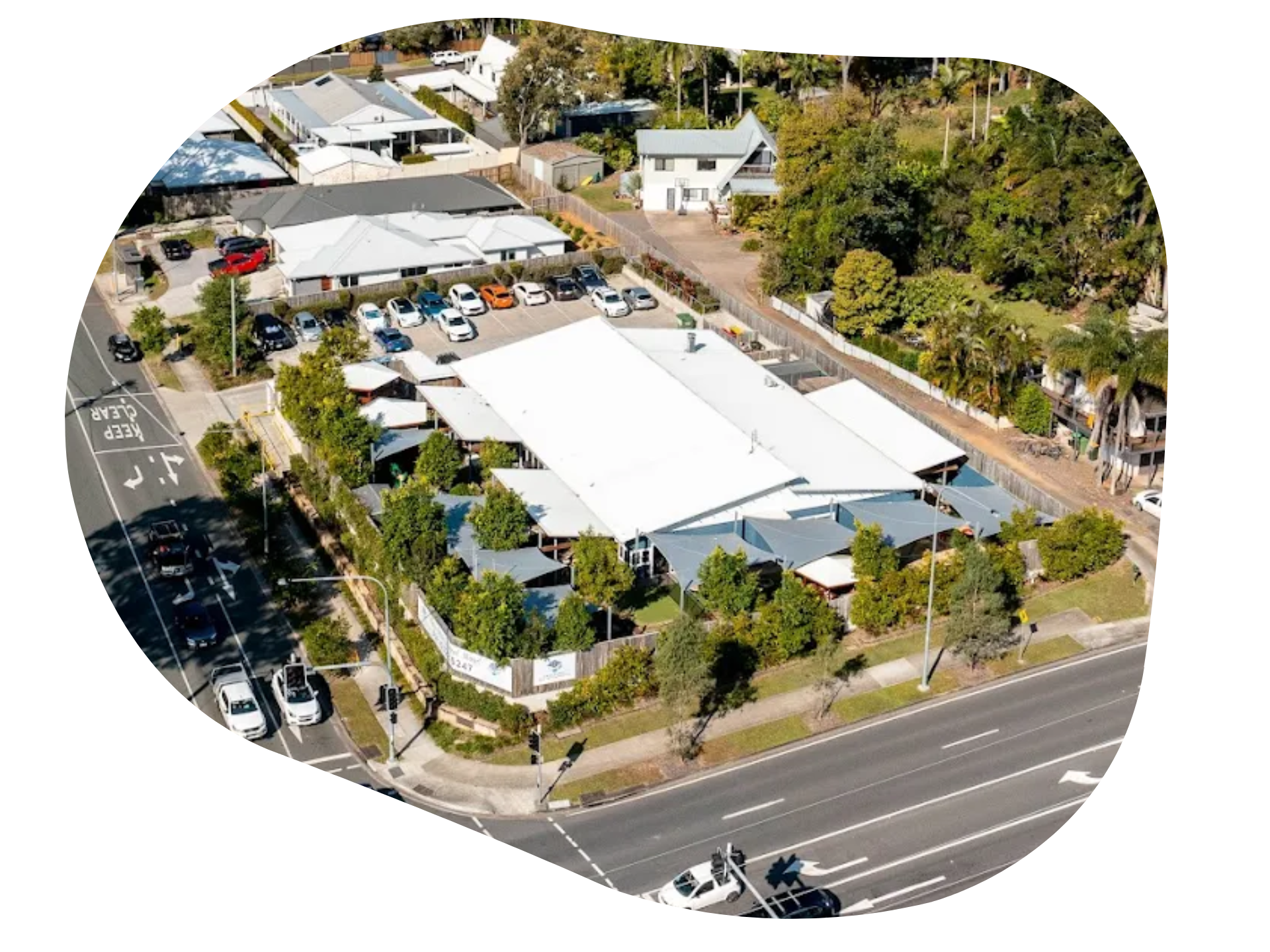 Aerial view of a commercial building with a white roof, surrounded by trees and a parking lot, near a road.
