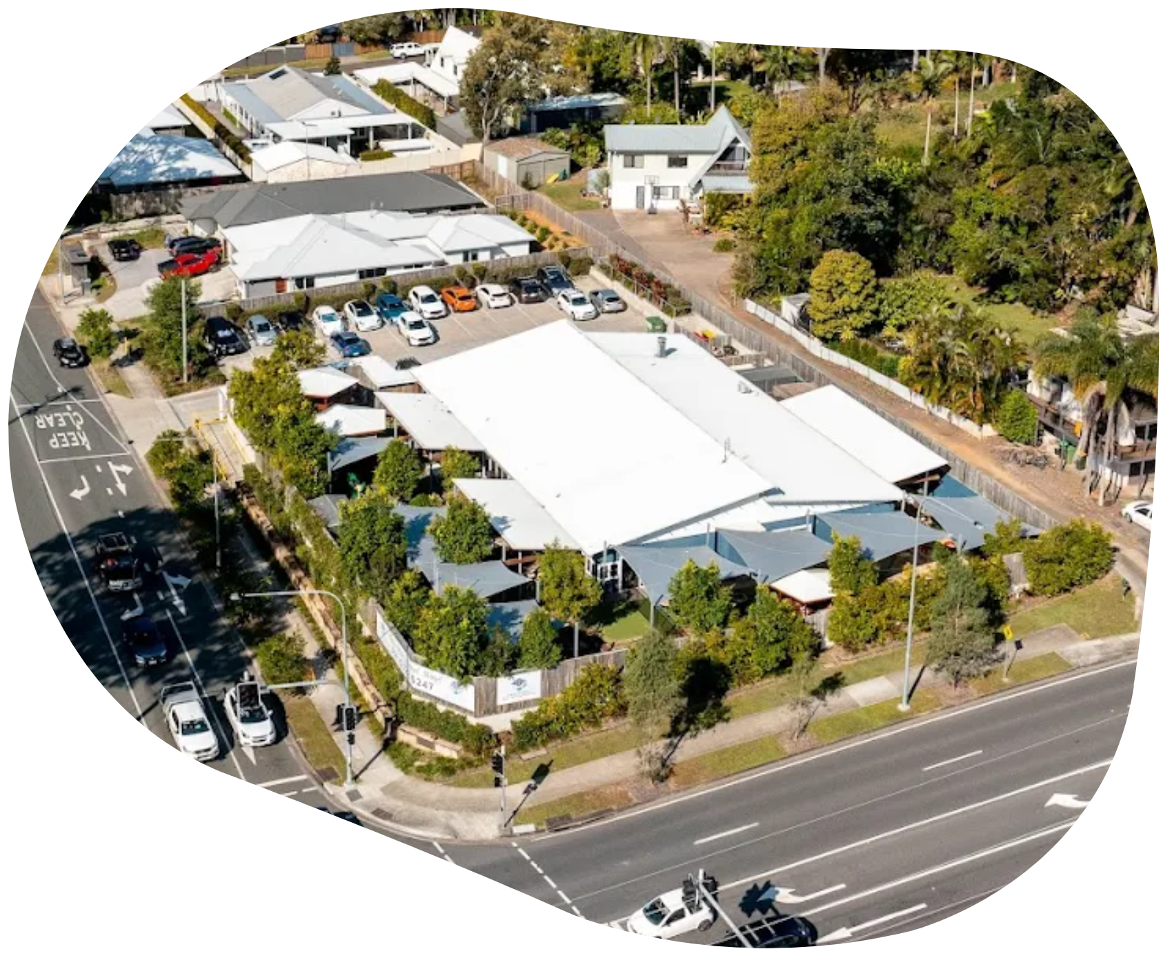 Aerial view of a white-roofed building and surrounding structures, with a parking lot and a road lined with cars and trees.