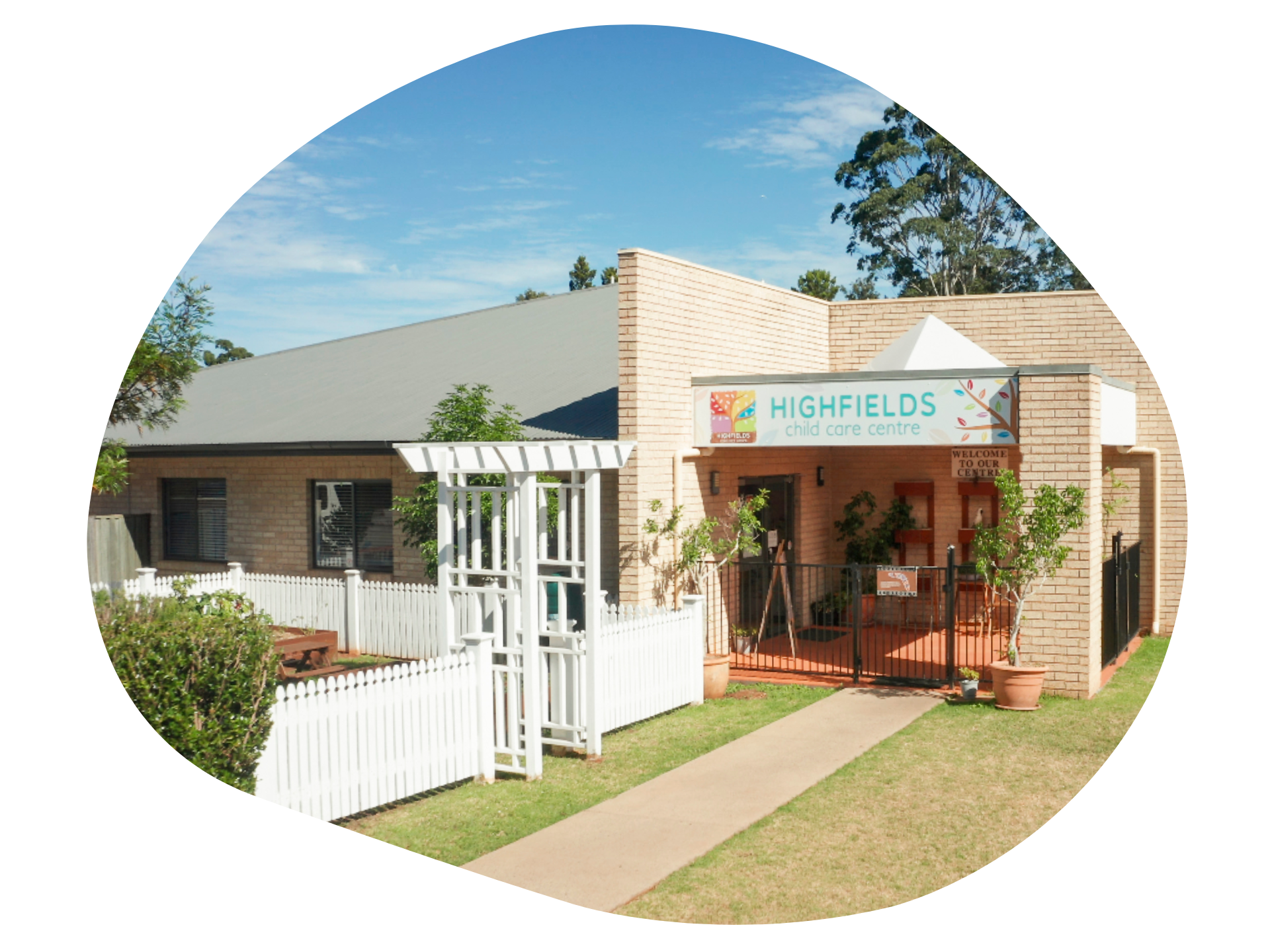 Highfields Early Learning Centre exterior, tan brick building, white picket fence, blue sky.