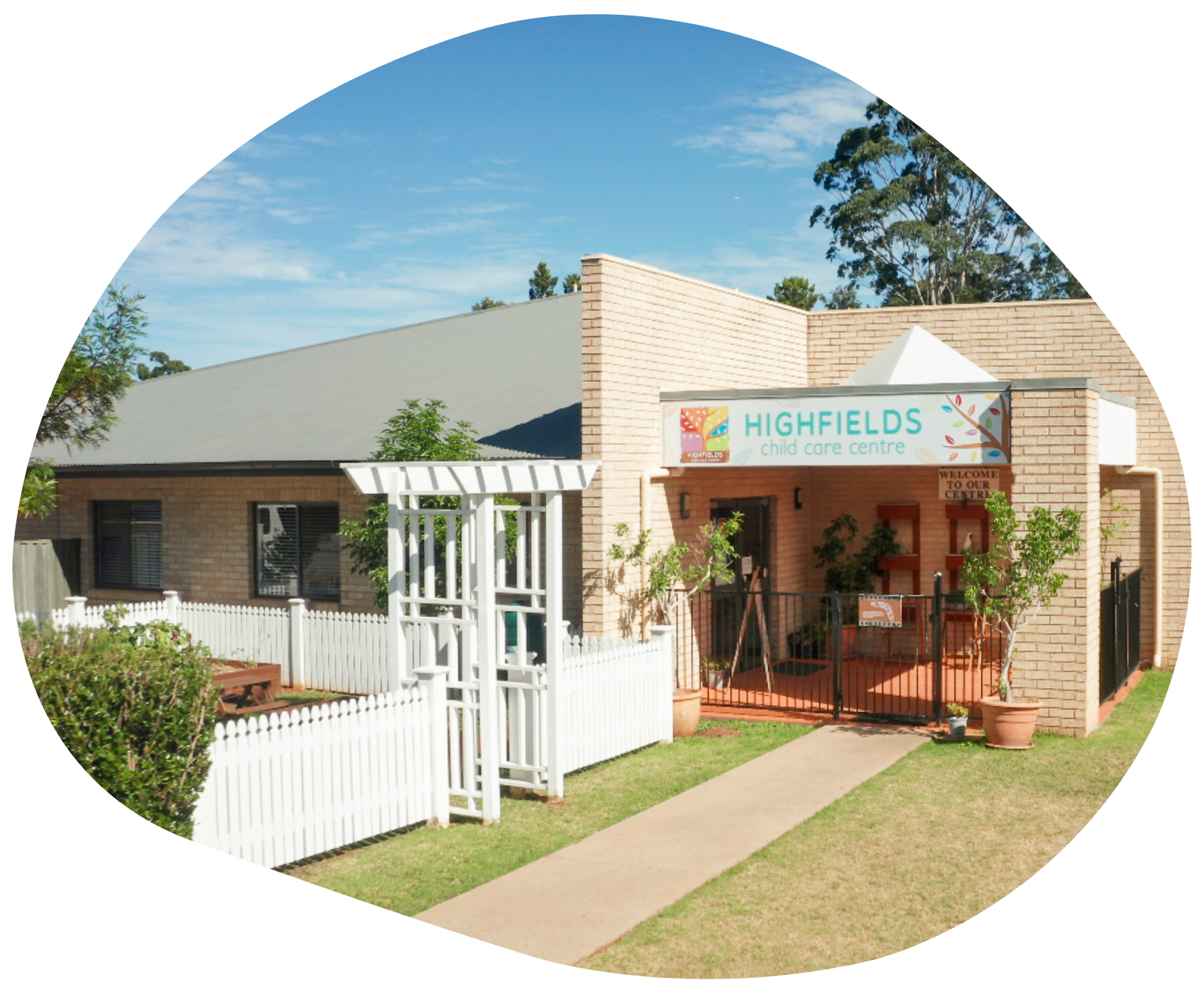 Highfields Early Learning Center, tan brick building with white picket fence, a garden, and blue sky.