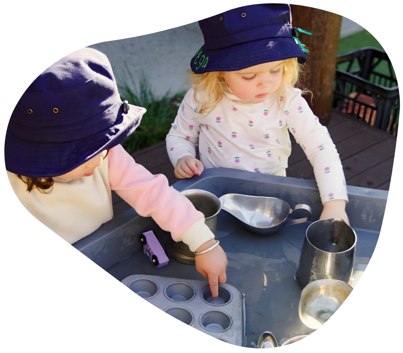 Two children in hats play with metal containers at a table outdoors.