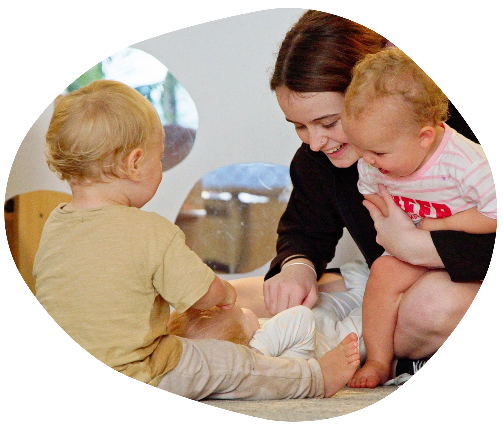 Woman smiles, changing infant's diaper with two toddlers nearby in a light-filled room.