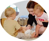 A caregiver changes a baby's diaper as two toddlers look on, all smiling and in a light-filled room.