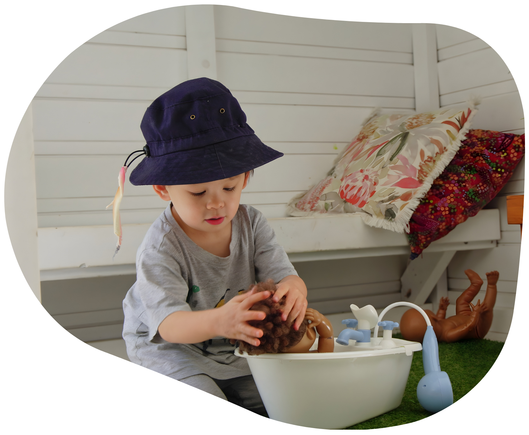 Child in a hat playing with a doll in a white basin, set against a white wood background.