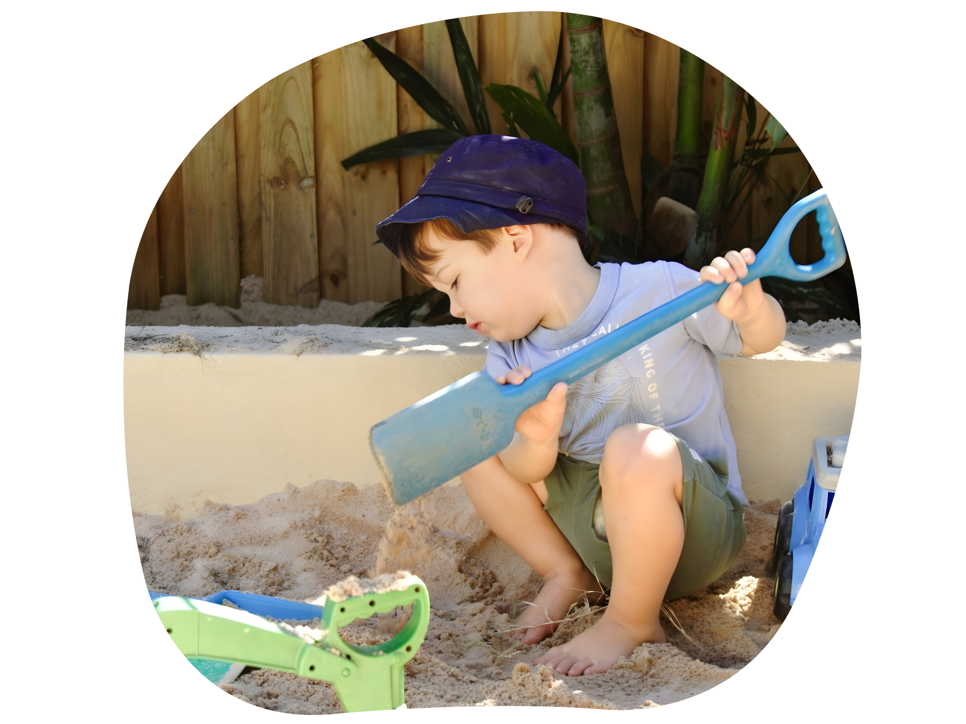 Boy in blue hat plays with sand and shovel in sandbox.