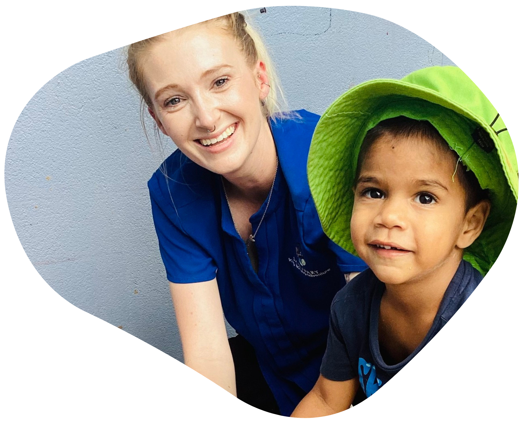 Woman in blue shirt smiles with a child in a green hat. They are close together indoors.