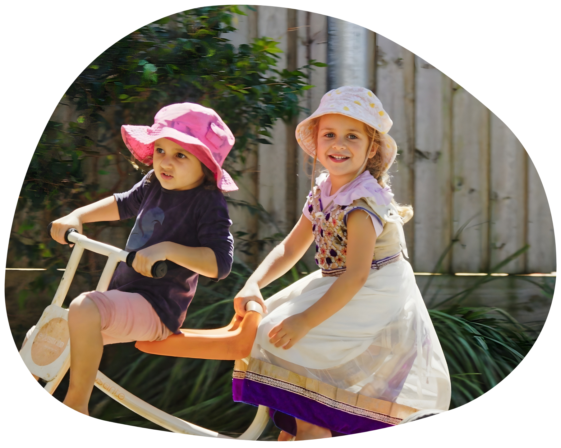 Two girls in hats ride a child's toy vehicle outdoors; one looks forward, one smiles.
