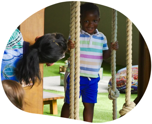 Boy smiling while holding rope in a play area, another child peeks around a wooden structure.