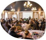 Conference room with presenter, audience seated at round tables, and a projection screen.