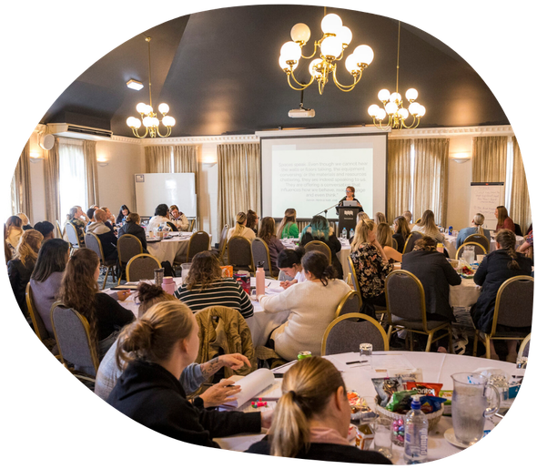 Large conference room; people seated at round tables, listening to a presentation on a screen.