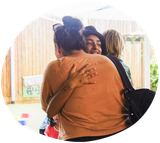 People embracing outdoors; one with a bun and sunglasses, another in a hat, smiling.