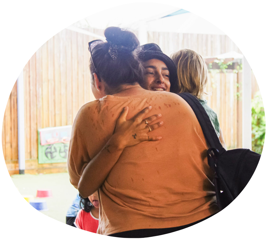 People hugging, smiling in front of a wooden fence; someone has a bag.