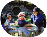 Children and teacher seated around a table, wearing blue hats. One child in a shark hat. Outdoor setting.