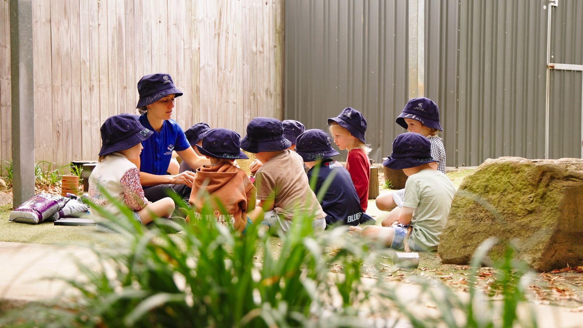 Children in blue hats sit with an adult, gathered outdoors.