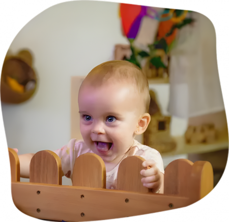 Smiling baby looking over a wooden fence, indoors, with toys in the background.