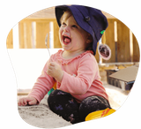 Child in pink shirt and blue hat, laughing while holding a spoon in a sandbox.