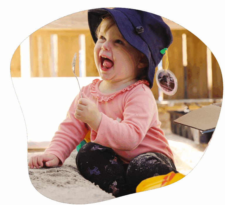 Child in pink shirt and blue hat, laughing while holding a spoon in a sandbox.