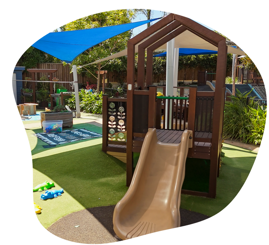 Playground with a brown slide, wooden structure, green turf, and blue shade.