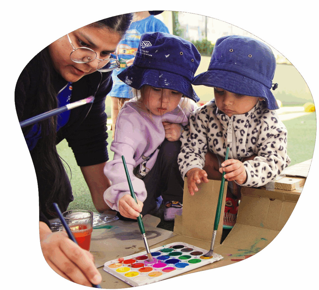 Children painting with guidance from a person, outdoors; colorful paint palette.