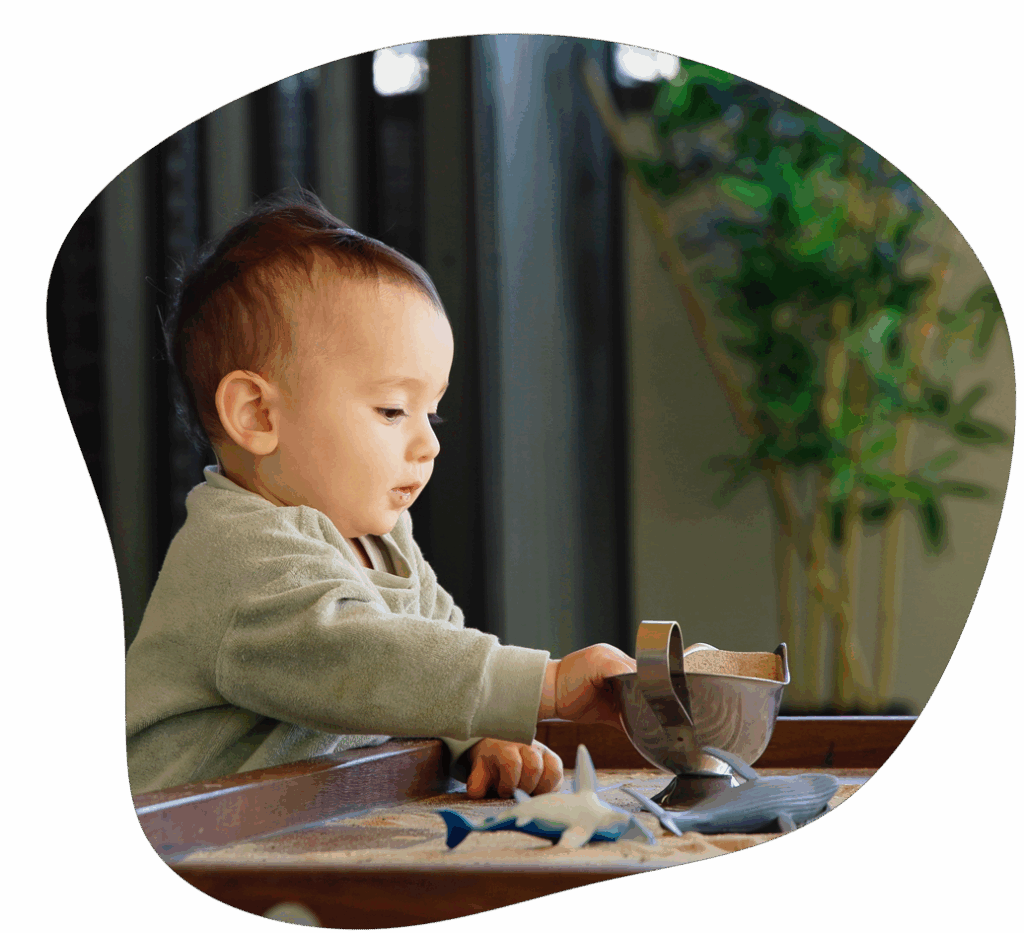 Baby reaching for an object on a tray; indoors, with a plant in the background.