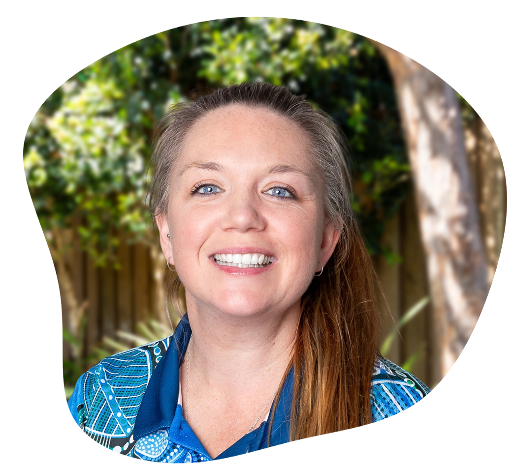 Woman with blue eyes and a smile, in a blue patterned shirt, with brown hair in a ponytail, outdoors.