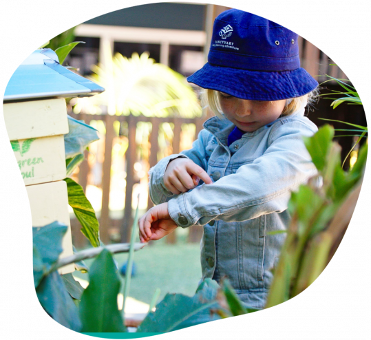 Young child smiles, holding flower, wearing a hat, red shirt, at a table with other flowers.