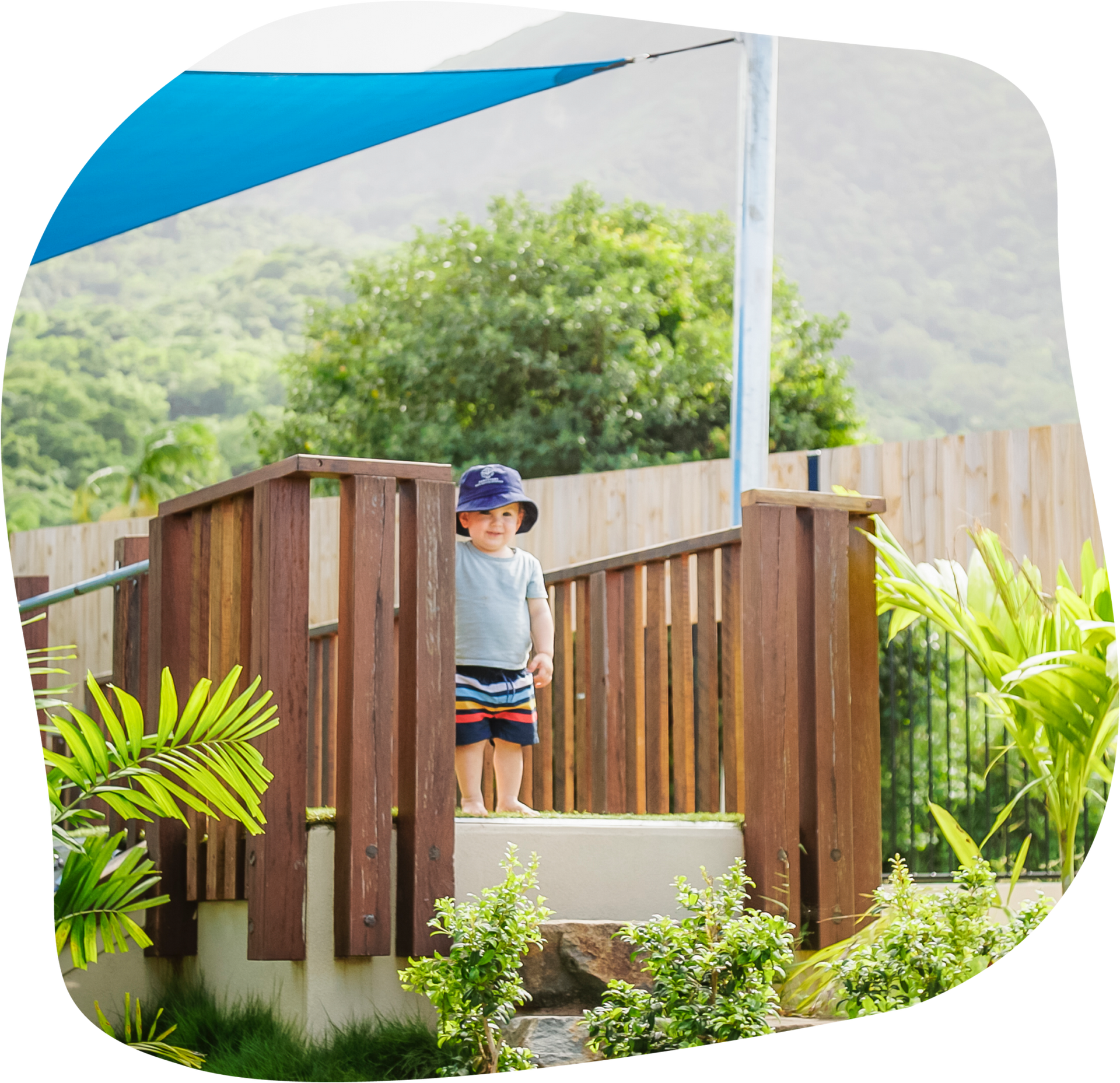 Child on a wooden deck, wearing a hat, under a blue shade sail. Green foliage and hills in background.