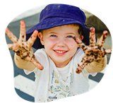 Smiling child with dirty hands wearing a blue hat, white shirt, outdoors.