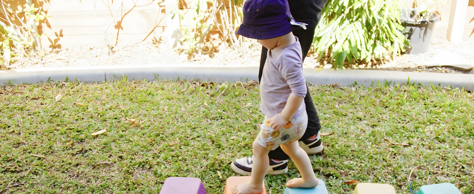 A child wearing a sun hat walks on colorful blocks in a grassy yard, with an adult nearby.