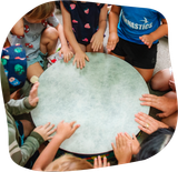 Children sitting in a circle, hands on a large, light-colored drum, indoors.