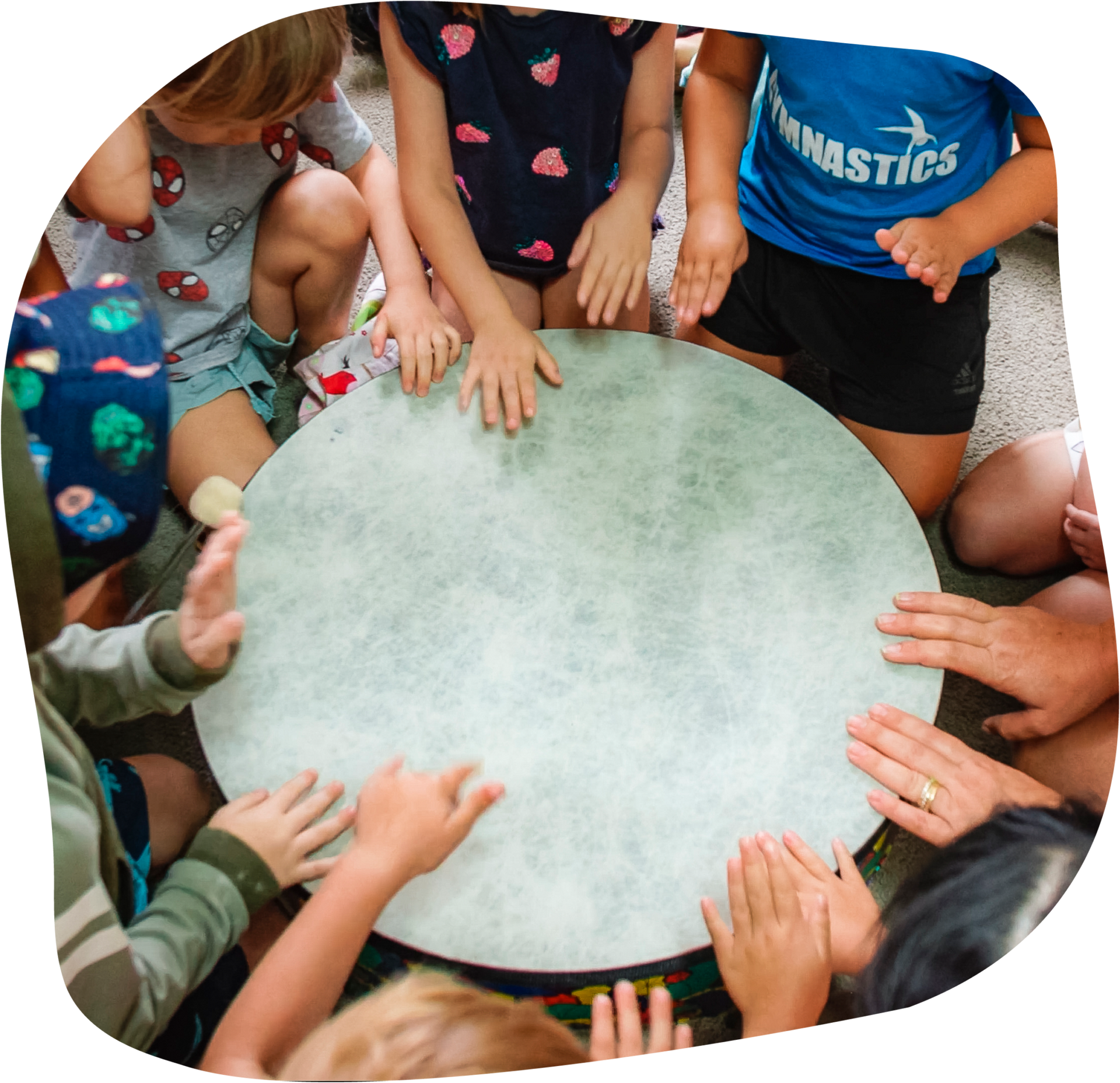 Children sitting in a circle, hands on a large, light-colored drum, indoors.