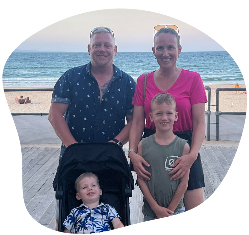 Family of four smiling on a boardwalk by the beach; one child in a stroller.