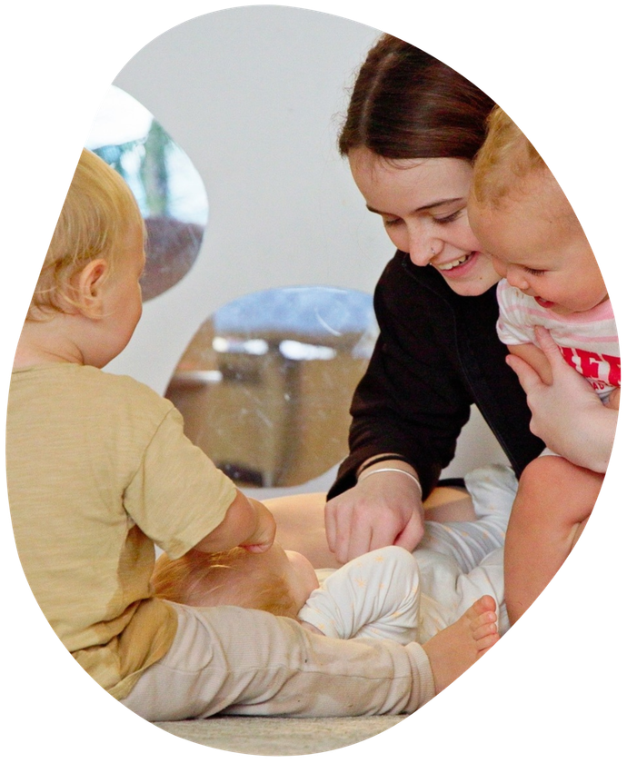 Two toddlers playing with a jar and spatula at a table indoors. One smiles.