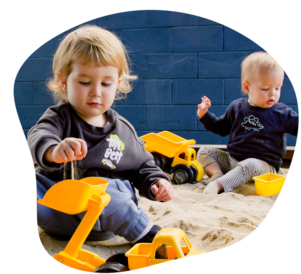 Two toddlers playing in a sandbox with yellow toy trucks and a shovel.